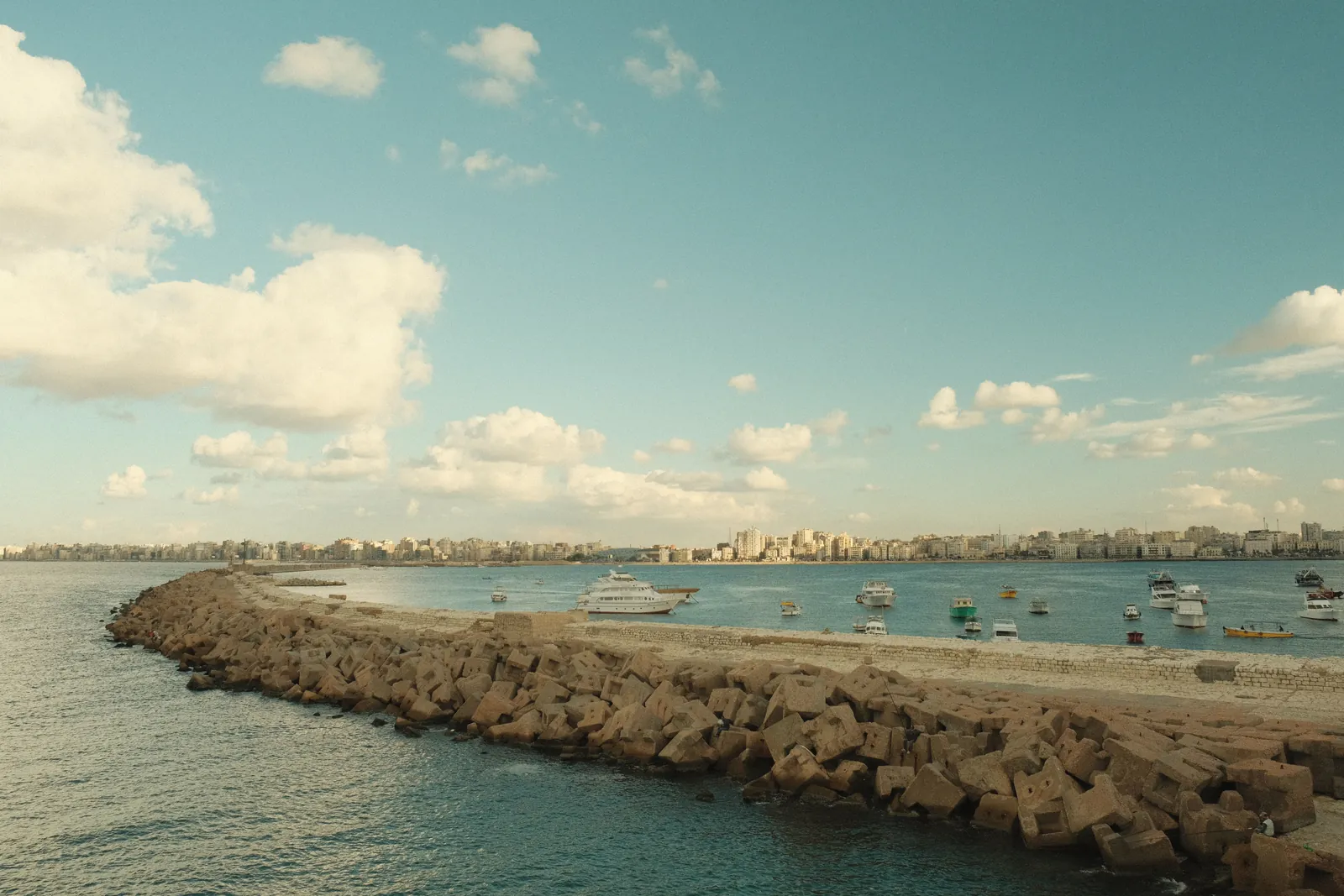 A picturesque view of a Mediterranean coastline with boats and distant city skyline under a partly cloudy sky.