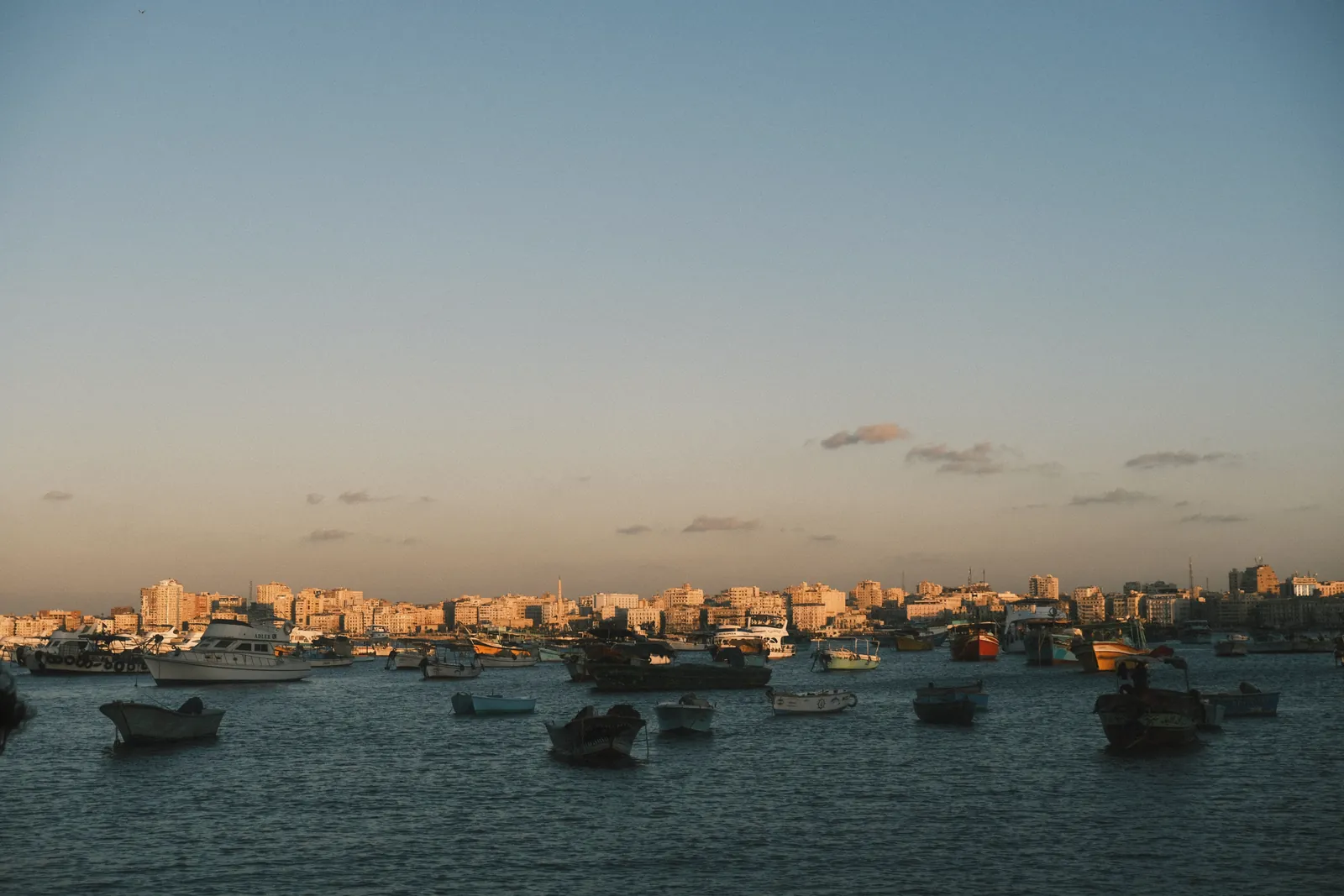 A serene view of boats in Alexandria's coastal harbor during sunset with city skyline.
