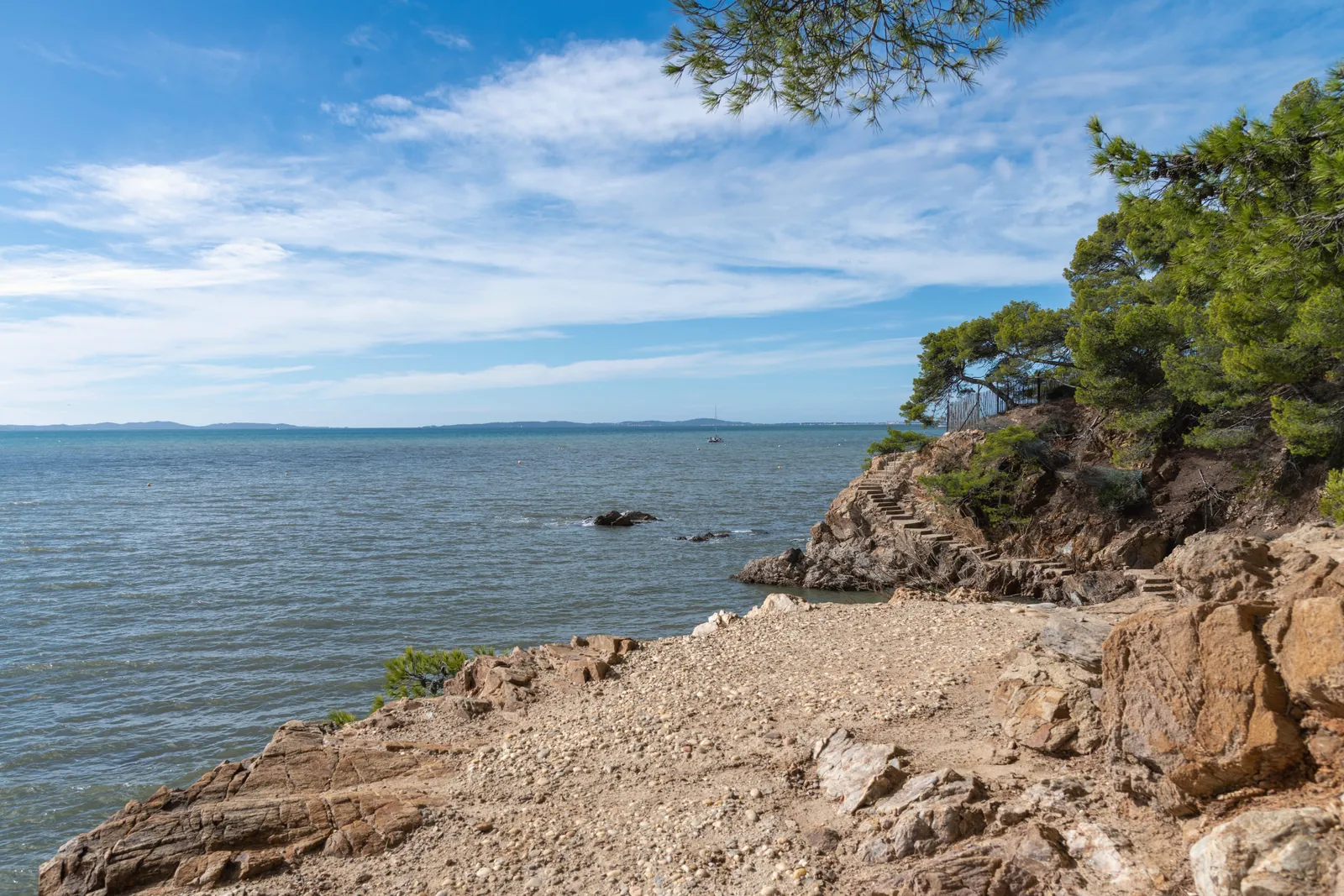 Peaceful view of a coastal rocky shore with stone steps and umbrella pines under a blue sky.