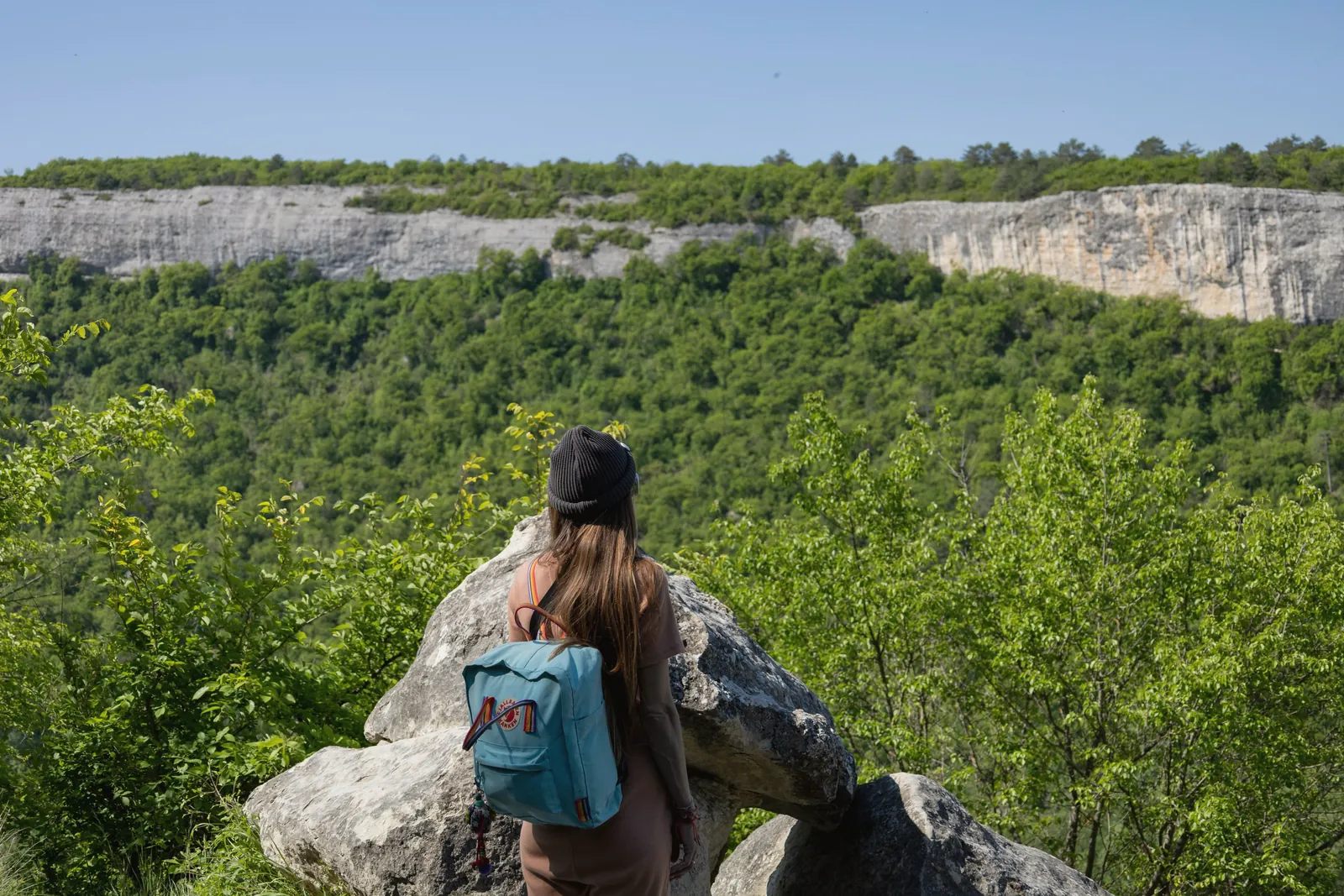 A hiker looks out over a lush green valley and rocky cliffs, embracing nature's beauty.