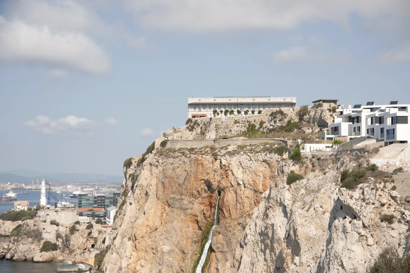 A stunning view of modern buildings atop a rocky cliff, overlooking a vibrant coastal city and harbor.