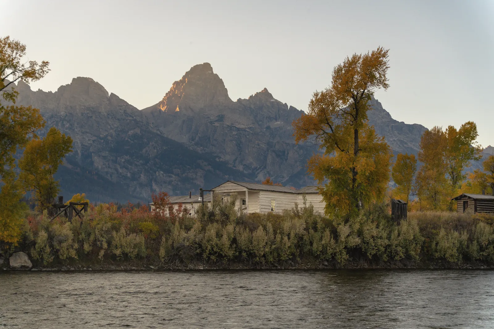 Beautiful autumn scene with a cabin by the river, set against the Grand Teton mountain range in Wyoming.