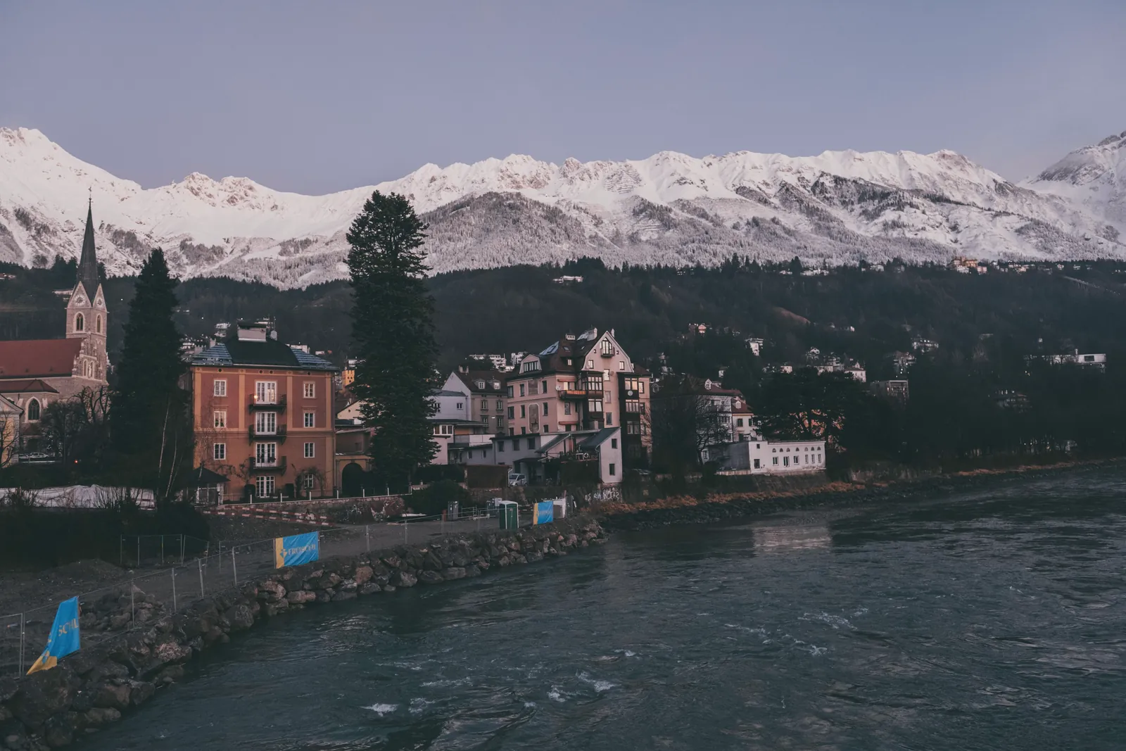 Charming Austrian town nestled by a river and snowcapped mountains at dusk.