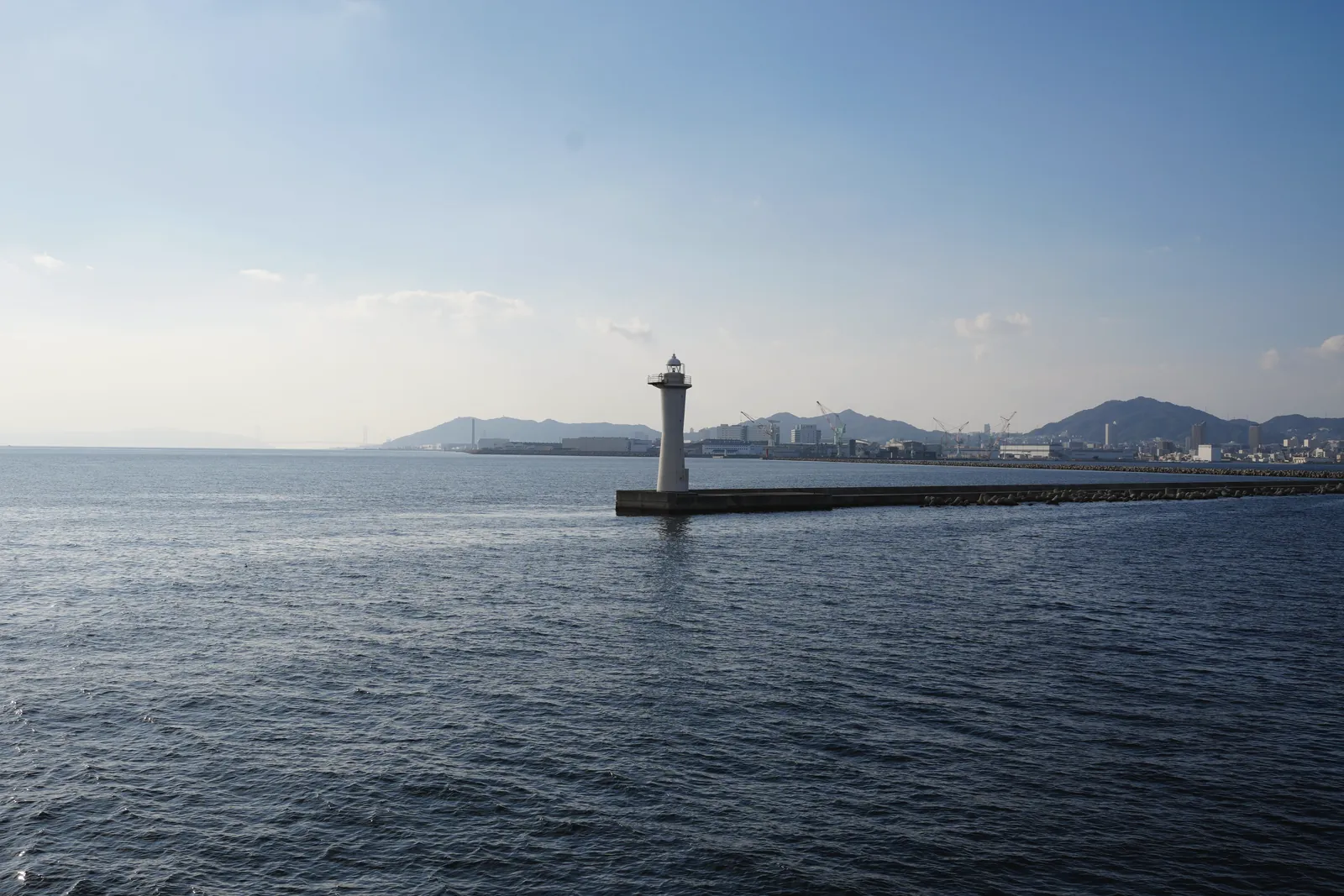 A tranquil view of a lighthouse against a backdrop of mountains and cityscape in Kobe, Japan.