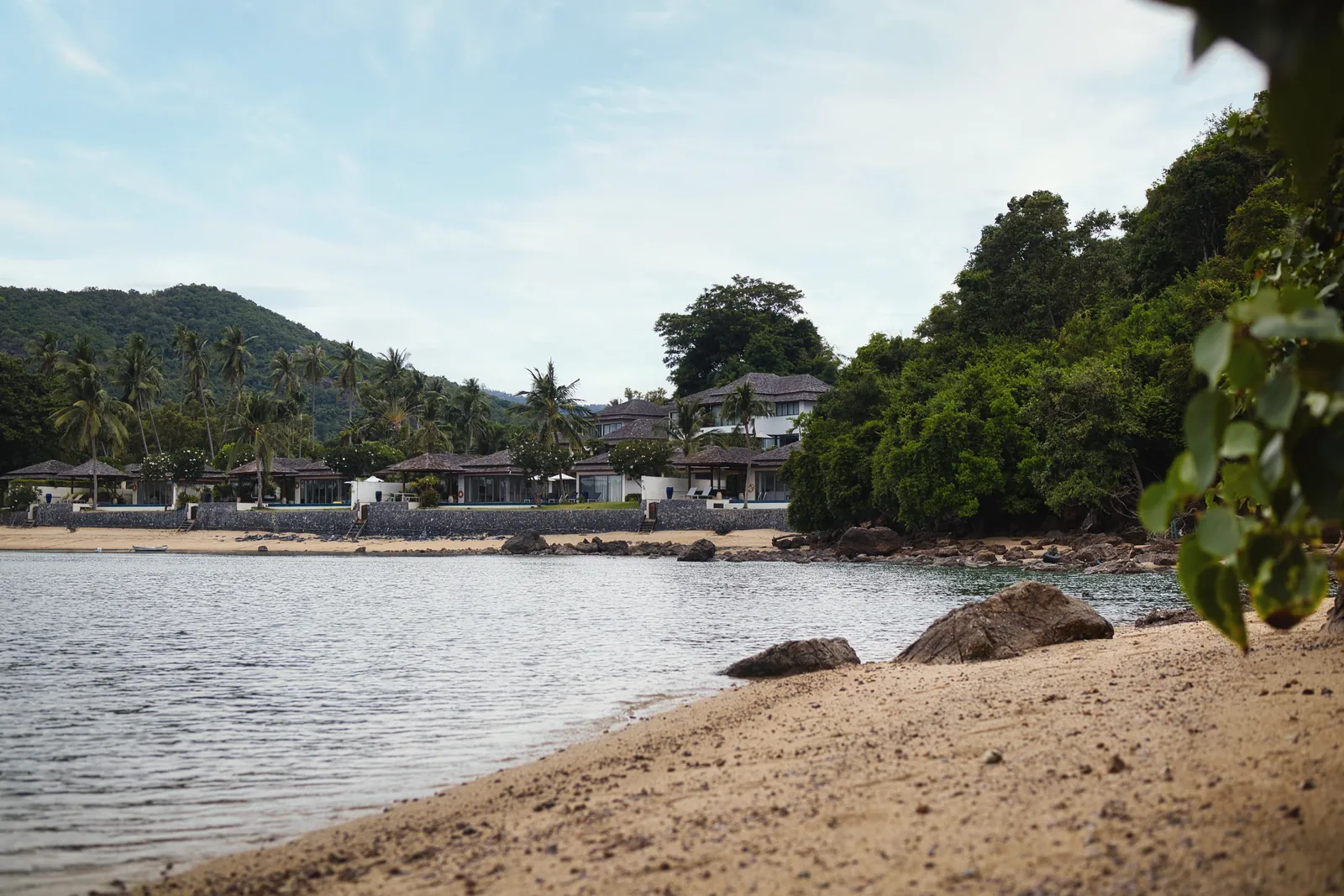 A peaceful tropical beach scene with lush greenery and distant villas under a blue sky.