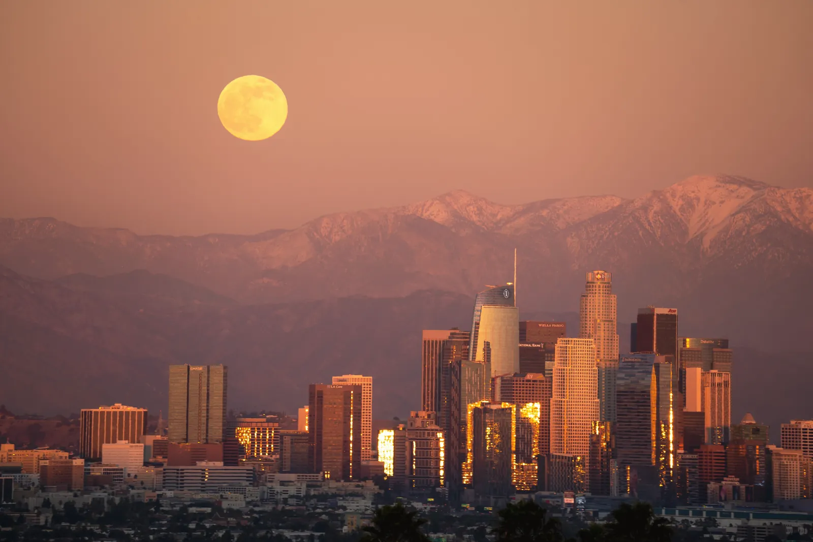 Stunning view of the Los Angeles skyline with a full moon rising over skyscrapers at sunset.