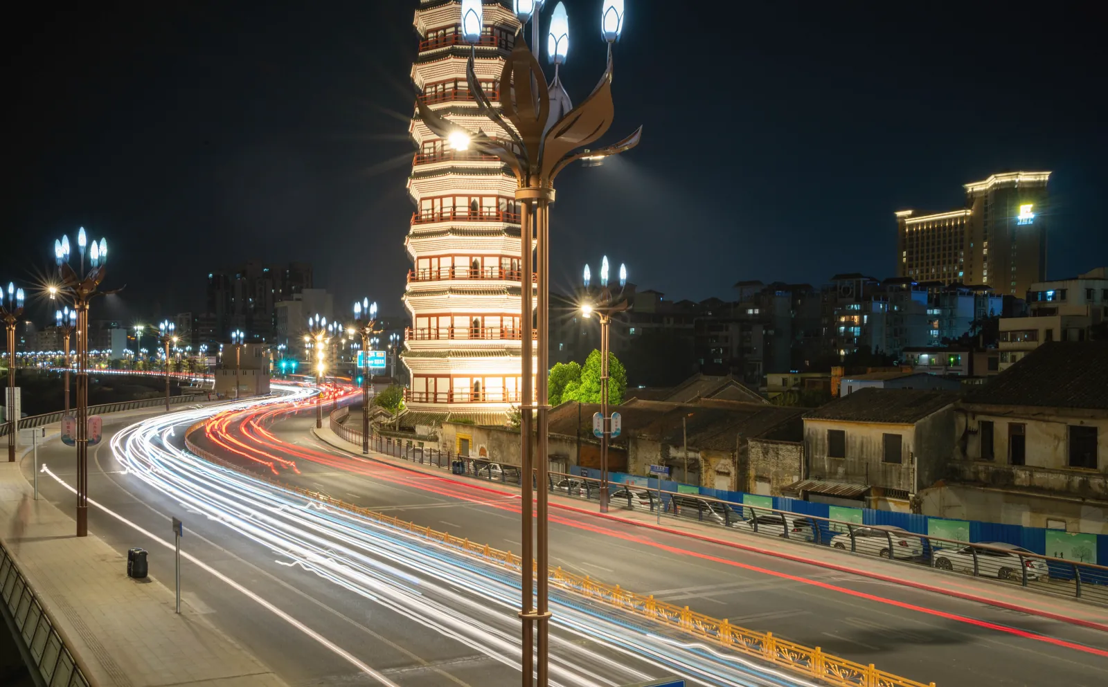 Vibrant cityscape featuring a historic pagoda lit up at night with dynamic light trails from passing vehicles.