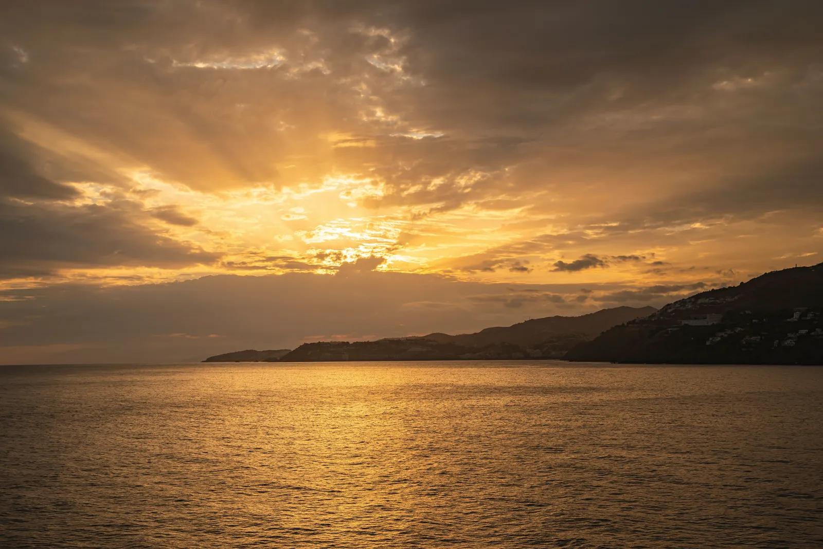 A breathtaking golden sunset casting radiant beams over the ocean horizon with silhouetted coastal hills.