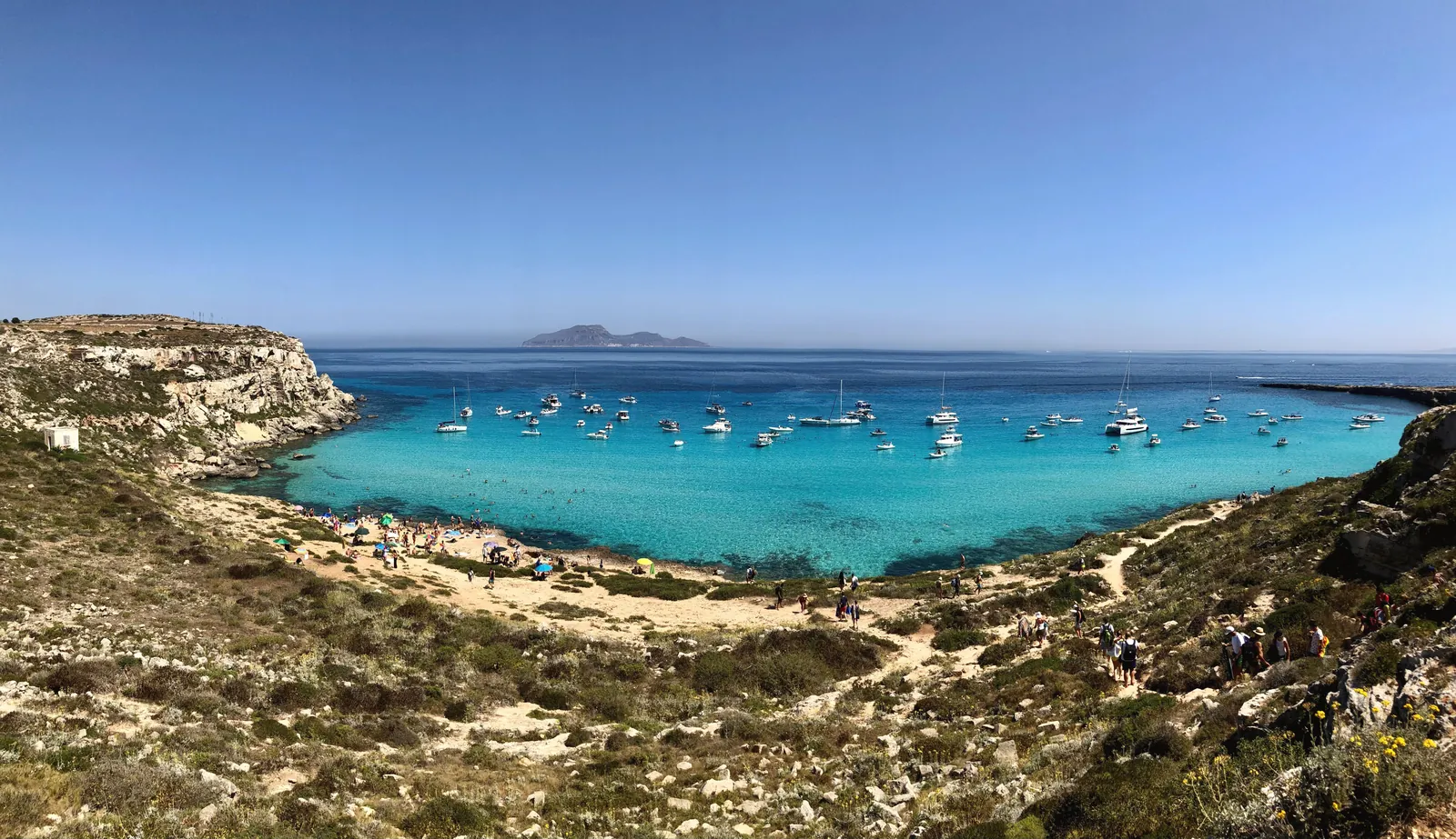 Beautiful bay with boats anchored in clear blue waters, surrounded by rocky cliffs and sunny skies.