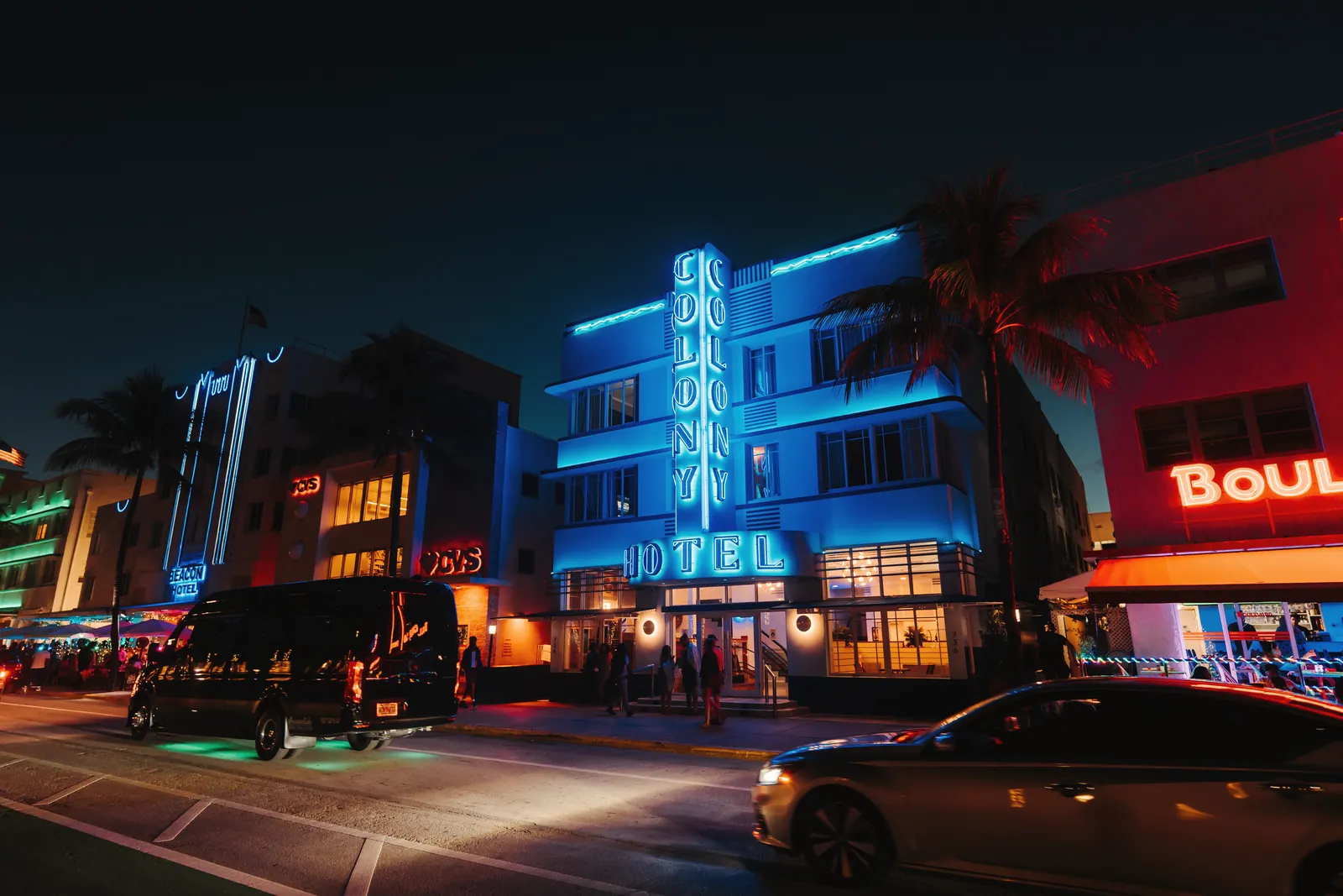 Glowing neon signs illuminate the Colony Hotel at night, showcasing vibrant nightlife in an Art Deco district.