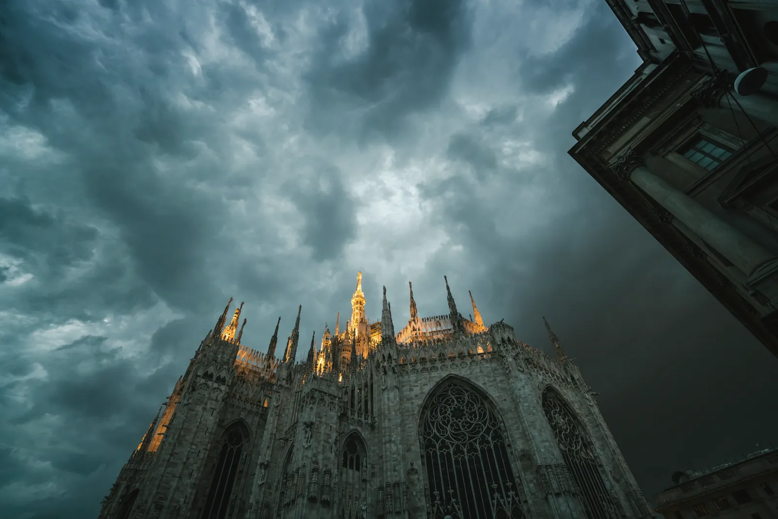 Low angle of Metropolitan Cathedral Basilica of Nativity of Saint Mary in Milan with sharp spires lit up with bright light against gloomy overcast sky