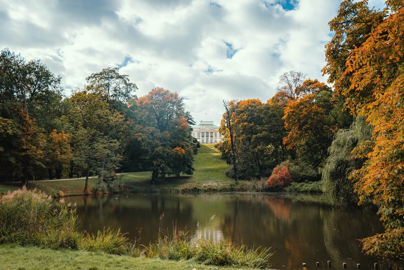 A grand mansion stands at the heart of an autumn landscape with vibrant foliage and a tranquil lake.