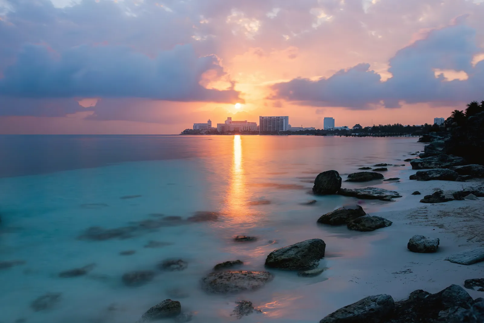 Serene sunrise over Cancun's rocky beach with a view of the Caribbean Sea and distant hotels.