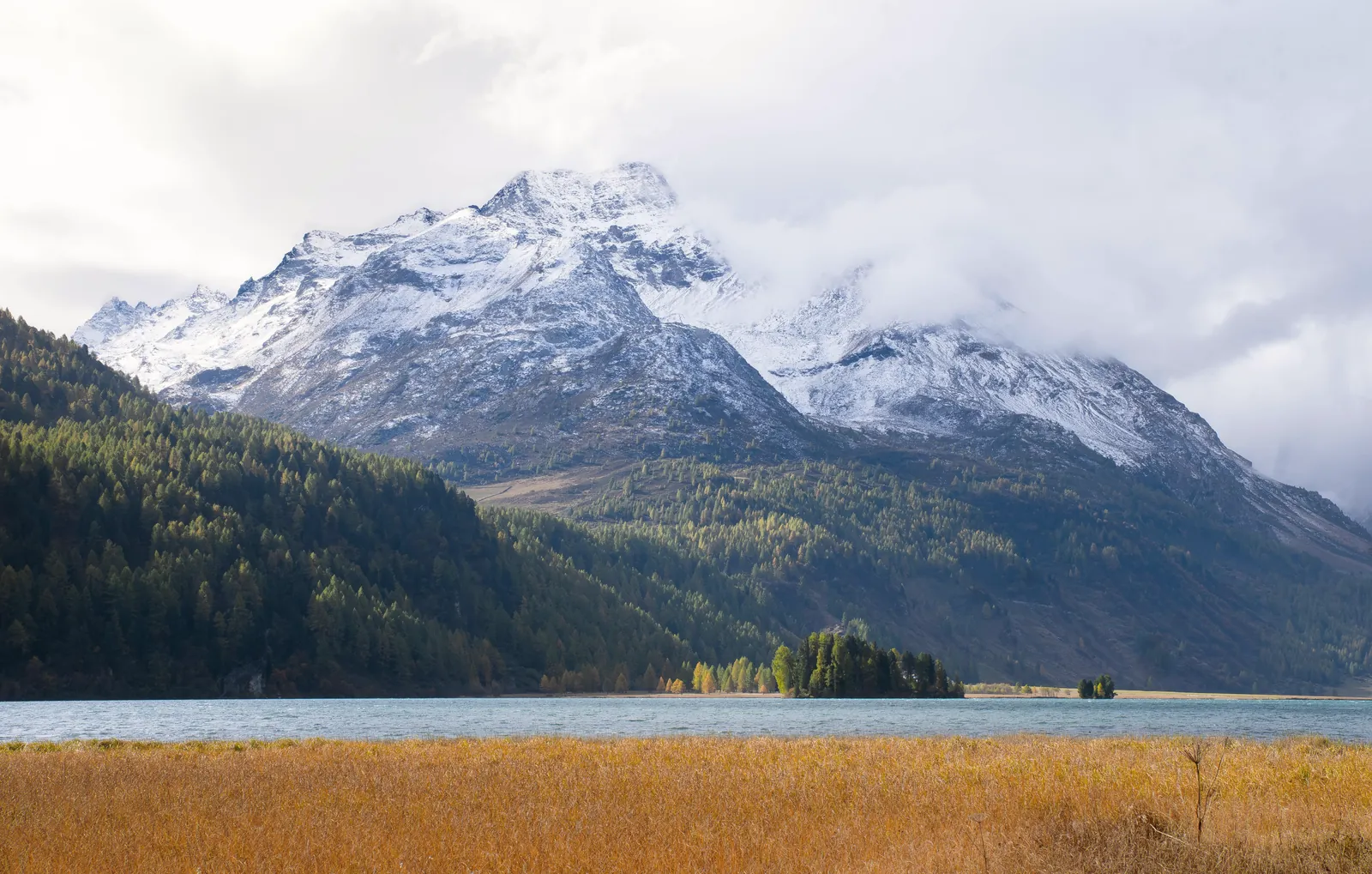 Breathtaking view of snow-capped alpine mountains, lush forests, and a serene lake in autumn.