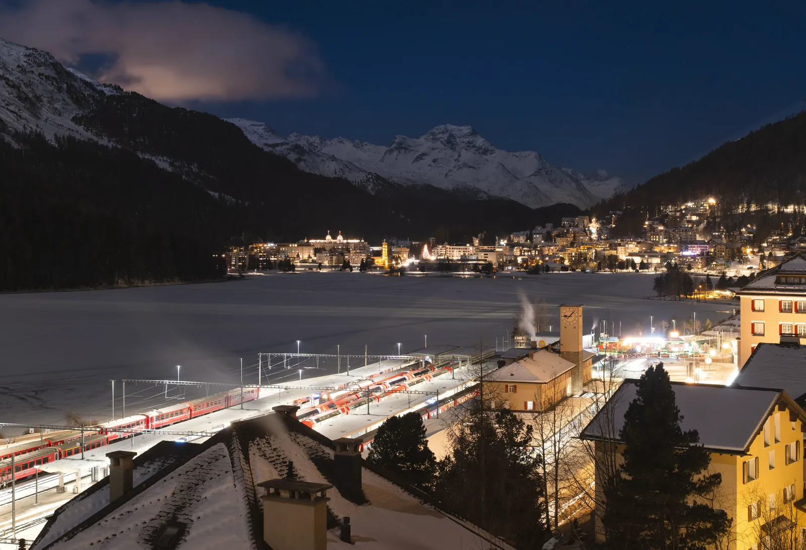 Scenic nighttime view over St. Moritz with illuminated train station and snowy mountains.