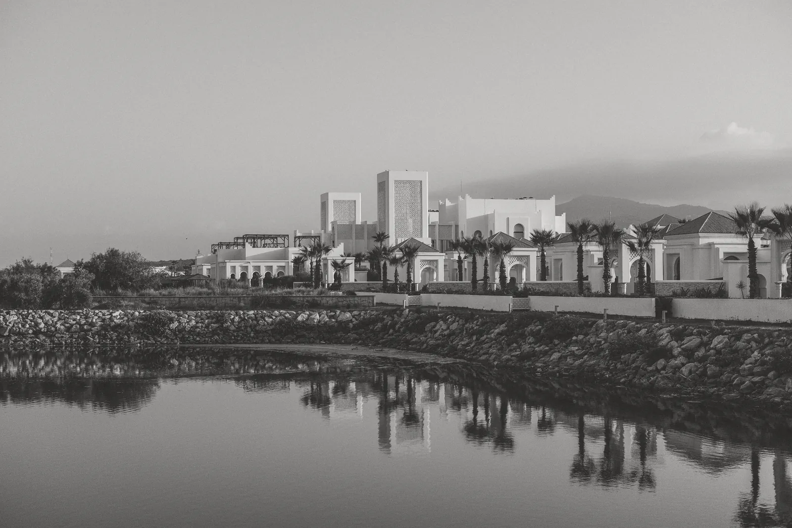 Black and white photo of elegant buildings and palm trees reflecting on water in Fnideq, Morocco.