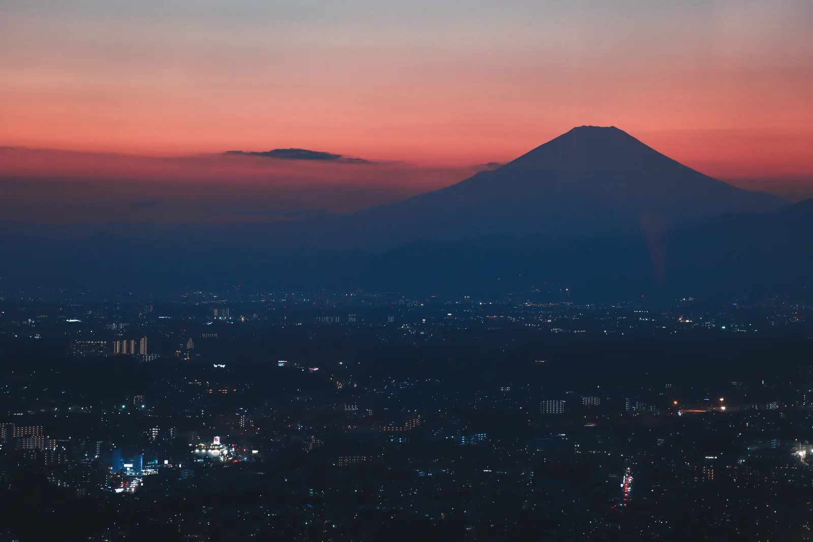 Mount Fuji silhouetted against a vibrant twilight sky over a bustling cityscape.