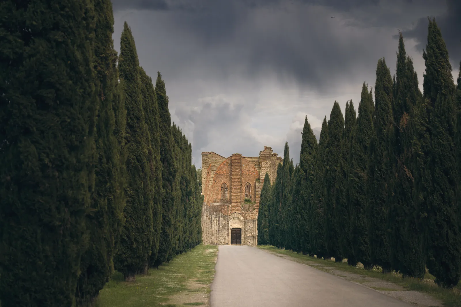 Dramatic view of San Galgano Abbey, Tuscany lined by trees with storm clouds overhead.