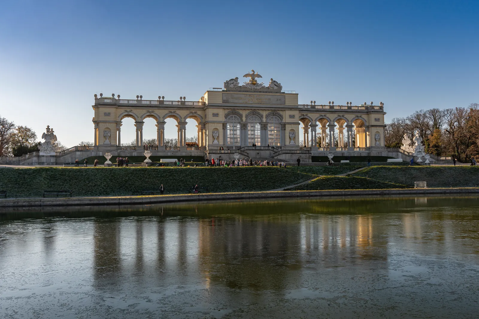 View of the Gloriette in Schönbrunn Palace, Vienna, reflecting on a pond under a clear blue sky.
